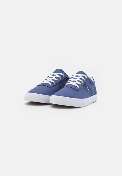 Tenis de lona azul-marinho com atacadores brancos, sola de borracha branca plana e ilhós em tom dourado. Apresenta um logotipo lateral e um design simples e minimalista.