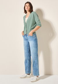 Lichtgroen geribbeld cardigan met een diepe V-hals, gecombineerd met hooglopende, vervaagde blauwe wijde jeans en witte sneakers met grijze accenten.