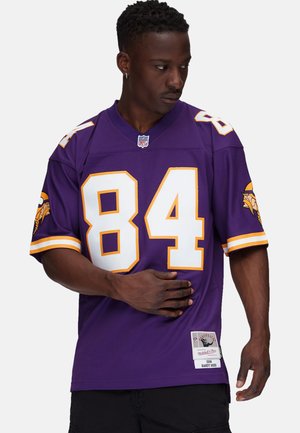 Man wearing purple NFL Vikings jersey number 84, standing with one hand on his stomach, looking down to his left.
