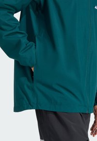 Veste teal avec une texture lisse, dotée d'une fermeture éclair et de poches latérales. L'ourlet est légèrement arrondi, et elle est associée à un pantalon sombre.