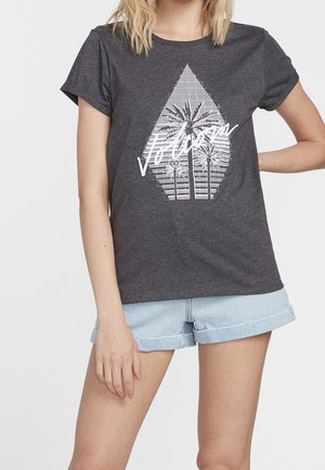 Nainen, jolla on päällään tummanharmaa T-paita, jossa on valkoinen geometrinen palmuaihe ja "Volcom"-teksti, yhdistettynä vaaleansiniset, käännetyt farkkushortsit.