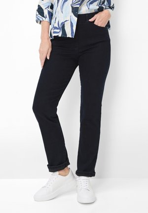 Woman wearing black slim-fit pants cuffed at ankles, paired with white sneakers and a blue patterned top, posing against a white background.