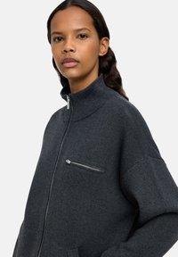 Młoda kobieta z ciemnymi włosami nosi ciemnoszary sweter zapinany na zamek z wysokim kołnierzem i zamykaną kieszenią na klatce piersiowej, stojąc na jednolitym tle.