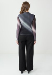 Kobieta z długimi falującymi włosami nosi dopasowaną bluzkę z długim rękawem z szarymi ukośnymi wzorami oraz czarne spodnie o szerokich nogawkach, stoi na tle jednolitego białego tła.