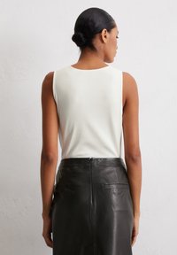 Weißes ärmelloses Oberteil, kombiniert mit einem hochgeschnittenen schwarzen Lederrock. Auffällige rückseitige Nähte und ein kleines Reißverschlussdetail an der Taille.