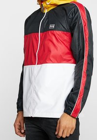 Mężczyzna w kurtce przeciwwietrznej Levi’s z czarnymi, czerwonymi i białymi blokami kolorów oraz żółtym kapturem, stojący z rękami opuszczonymi wzdłuż ciała.