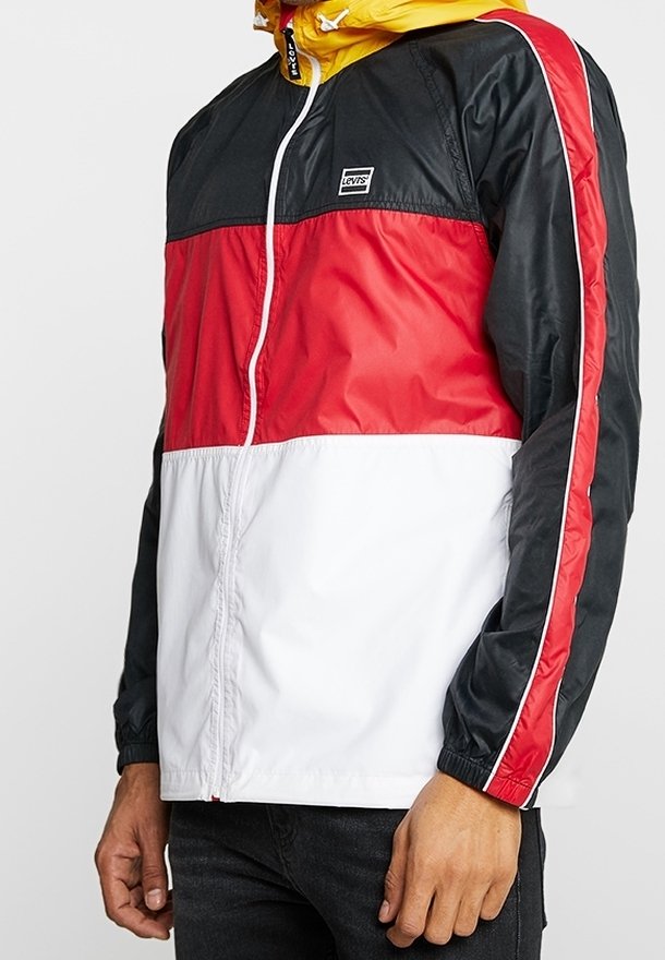 Mężczyzna w kurtce przeciwwietrznej Levi’s z czarnymi, czerwonymi i białymi blokami kolorów oraz żółtym kapturem, stojący z rękami opuszczonymi wzdłuż ciała.