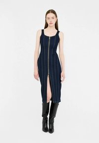 Robe en denim foncé avec une fermeture éclair à l'avant, une fente jusqu'au genou et des surpiqûres contrastantes. Le mannequin porte des bottes noires montant jusqu'au genou. Design simple et ajusté.