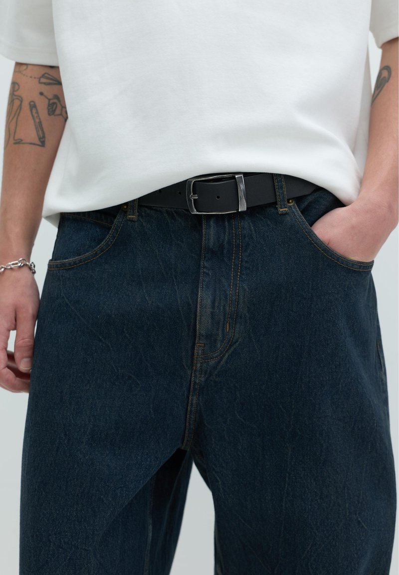 Personne portant un jean en denim foncé avec les mains dans les poches, une chemise blanche rentrée et une ceinture noire avec une boucle rectangulaire argentée.
