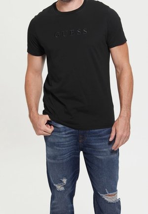 Mand iført sort Guess T-shirt og slidte blå jeans, stående med den ene hånd i lommen mod en ensfarvet baggrund.