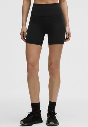 Donna che indossa pantaloncini da biker a vita alta neri, calzini neri e scarpe da ginnastica nere, in piedi contro uno sfondo chiaro uniforme.