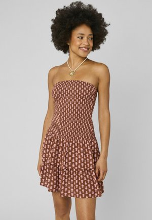 Femme aux cheveux bouclés portant une robe marron bustier avec un motif rose et un collier en forme de soleil, souriante et regardant vers la droite.