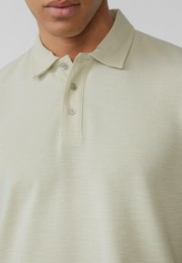 Vaaleanvihreä polo-paita, joka on valmistettu teksturoidusta kankaasta ja jossa on klassinen kaulus. Paidassa on kolme nappia ja hienovarainen kuviointi koko pinnalla.