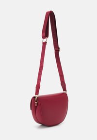 Červená kožená kabelka přes tělo s křivkovým tvarem, označeným popruhem a zlatými kovovými detaily. Popruh obsahuje vyražené logo.