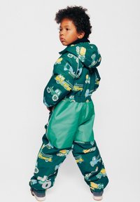 Mono impermeable verde con estampados coloridos de vehículos de construcción. El diseño incluye una capucha, puños elásticos y un ajuste ceñido para mantener el calor.
