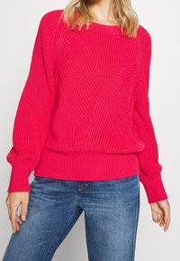 Femme portant un pull en maille texturé rose vif avec des manches bouffantes et un jean en denim bleu sur un fond uni.