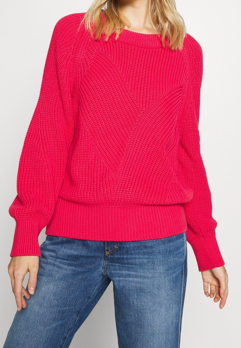 Femme portant un pull en maille texturé rose vif avec des manches bouffantes et un jean en denim bleu sur un fond uni.