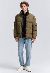 Olivově zelená prošívaná péřová bunda s límcem stojáčkem a zipem, nošená přes námořnicky modrou mikinu, doplněná světle modrými džíny a bílými teniskami.