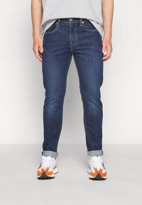 Tmavě modré džíny s úzkým střihem, s oranžovým švem, pasem s poutky na opasek a manžetami vyhrnutými nad kotníky.