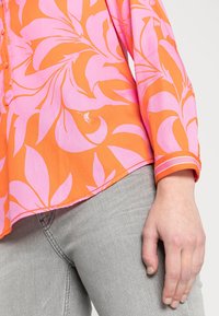 Blouse orange à manches courtes avec motif de feuilles roses, boutonnée sur le devant, et manches à revers rayés contrastés. Associée à un jean denim gris clair.