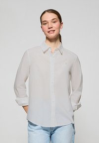 Biała koszula z długim rękawem, wykonana z lekkiego materiału, z kołnierzykiem i małym haftowanym logo na piersi.