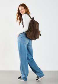 Sac à dos en velours côtelé marron avec des détails plissés, deux poches avant et des bretelles réglables. Associé à un jean ample bleu clair et des baskets noires.