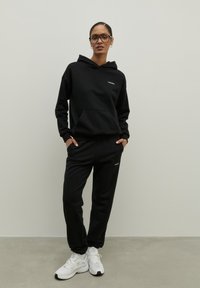 Čierna mikina s kapucňou a predným vreckom, malým logom, v kombinácii s čiernymi teplákmi, obe vyrobené z mäkkého materiálu. Celkový vzhľad dopĺňajú biele športové tenisky.