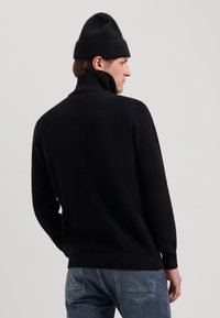 Maglione nero lavorato a maglia con colletto alto, texture a coste e maniche lunghe, abbinato a un berretto nero coordinato. Vista da dietro.