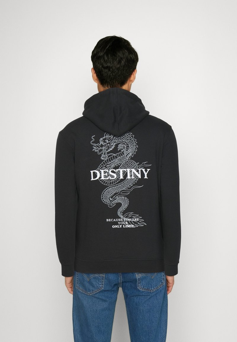 Hombre con sudadera negra con gráfico de dragón blanco y el texto "DESTINO" y "Porque tú eres tu único límite" en la parte de atrás, combinado con vaqueros azules.