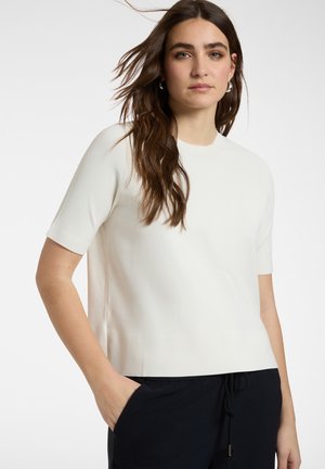 Frau mit langen braunen Haaren, die ein weißes kurzärmeliges Oberteil und schwarze Hose trägt, steht mit einer Hand in der Tasche vor einem schlichten Hintergrund.