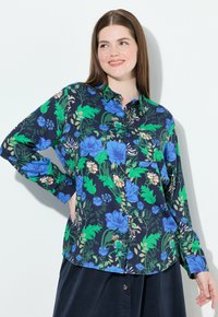 Donna con capelli castani che indossa una camicia blu navy con bottoni, decorata con un motivo floreale verde e blu, in piedi davanti a uno sfondo bianco, sorridente.
