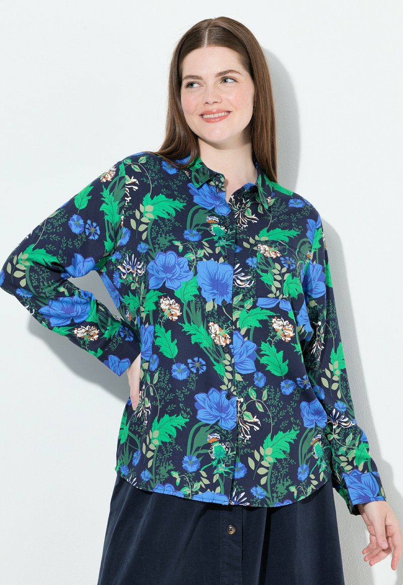 Donna con capelli castani che indossa una camicia blu navy con bottoni, decorata con un motivo floreale verde e blu, in piedi davanti a uno sfondo bianco, sorridente.