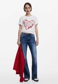Bílý crop top s červeným srdíčkem a textem "desigual", kombinovaný s rozšířenými modrými džíny. Modelka drží červenou bundu.