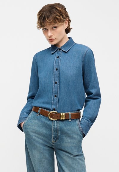 Άτομο που φοράει μπλε τζιν πουκάμισο με κουμπιά, μαζεμένο σε ψηλόμεσα τζιν με καφέ ζώνη, χέρια στις τσέπες, ουδέτερο φόντο.