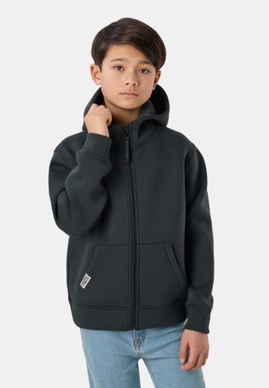 Dreng med mørkt hår iført en sort hættejakke med lynlås og lyseblå jeans, som holder ved kraven på hættejakken med den ene hånd.