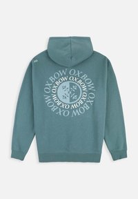 Sweat à capuche en teal, avec un logo circulaire blanc et du texte au dos. Manches et ourlet côtelés, matière douce avec une texture lisse.