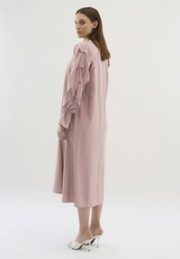 Rochie lungă, lejeră, dintr-un material roz moale, cu mâneci volănaș și manșete strânse. Modelul poartă pantofi cu vârf deschis albi.