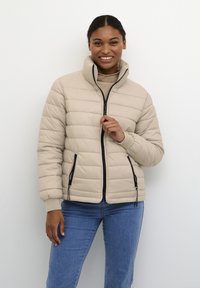 Jonge vrouw die glimlacht, gekleed in een beige gewatteerde jas boven een tan trui en blauwe spijkerbroek, staande tegen een witte muur, hand bij de rits van de jas.