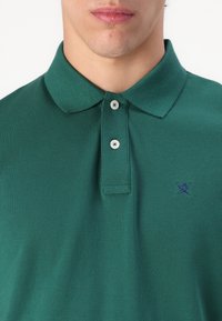 Tamsiai žalias polo marškinėlis, pagamintas iš tekstūruotos medžiagos, turintis apykaklę, tris sagas ir mažą mėlyną logotipą kairiajame krūtinės pusėje.