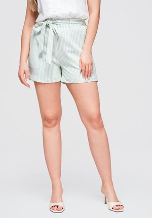 Femme portant un short rayé vert clair et blanc avec une ceinture nouée, un haut blanc sans manches et des talons blancs ouverts, debout devant un fond uni.