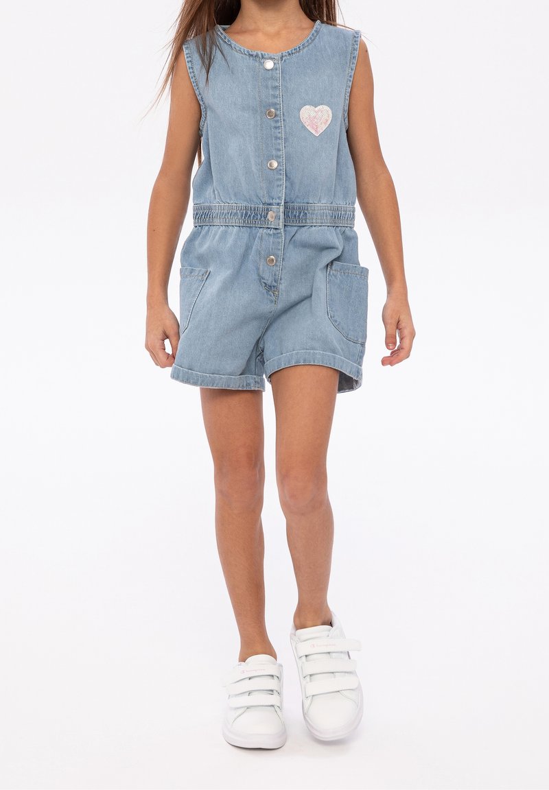 Enfant portant une combinaison courte en denim sans manches avec des boutons à l'avant et un patch en forme de cœur rose, associée à des baskets blanches à scratch.