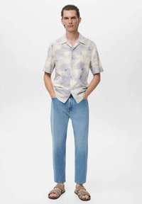 Kurzärmliges, hellfarbiges Button-Up-Hemd mit Wolkenmuster, kombiniert mit hellblauer Jeans und braunen Riemchensandalen, Hände in den Taschen.