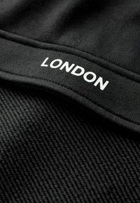 Tela negra con el texto blanco "LONDRES" impreso sobre una superficie textil ribeteada doblada, mostrando detalles de textura y costuras.