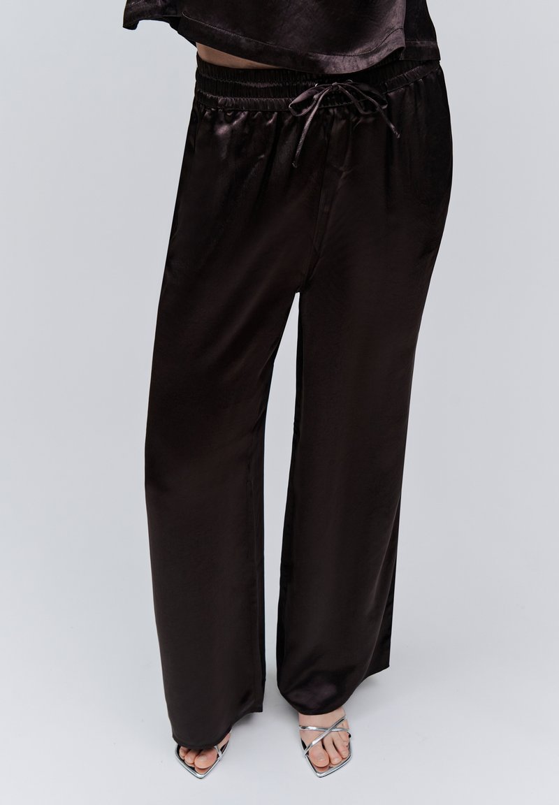 Pantalon large en satin noir avec taille à cordon porté avec des talons ouverts argentés à lanières sur un fond neutre.