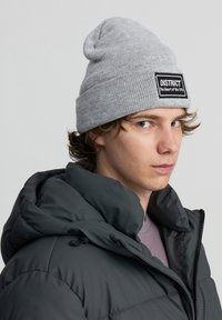 Beanie grigia a maglia con orlo rollato e un'etichetta rettangolare nera con la scritta "DISTRICT Il cuore della città", indossata sopra una giacca a bomber scura.