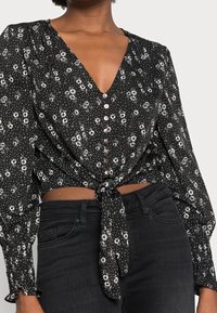 Haut portefeuille noir à motif floral avec un col en V, des manches longues bouffantes, des boutons sur le devant et un détail de nœud à la taille, associé à un jean noir.