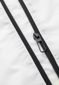 Curseur de fermeture éclair noir sur des dents de fermeture éclair noires, fixé à un tissu blanc, avec le logo "ombre" visible sur la tirette.