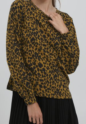 Kvinde iført brun og sort leopardmønstret langærmet top med plisseret sort nederdel, den ene hånd rører ved kraven.