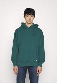 Sudadera de color verde azulado con un bolsillo frontal, capucha con cordón y puños de canalé. Presenta un pequeño logotipo negro en el pecho y una etiqueta en contraste en el dobladillo.