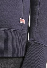 Mörkblå sweatshirt med ribbade ärmslut och nederkant, med en liten röd och vit logotags sydd på sidan. Mjuk, texturerad tygmaterial.